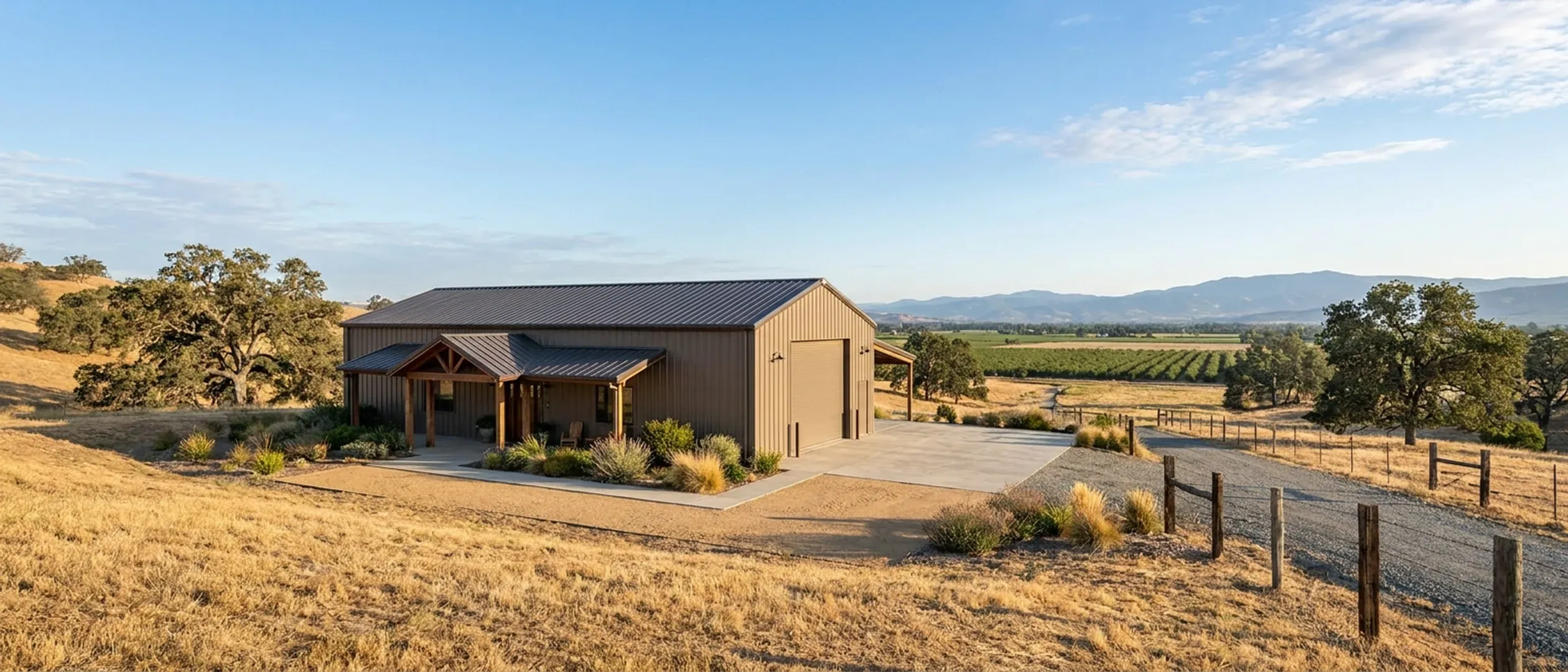 A completed barndominium with a covered entry porch and an attached shop bay, golden oak-dotted hills behind under a clear sky.