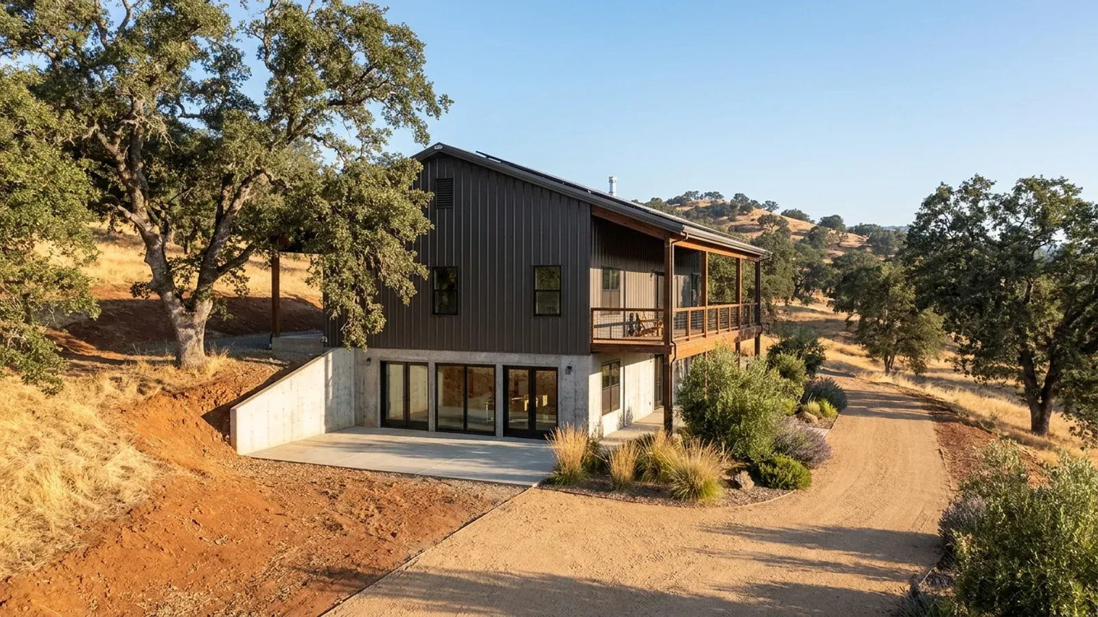 A barndominium with a walkout lower level built into a sloping lot, the lower level opening onto a patio at grade, with oaks on the golden hillside behind.